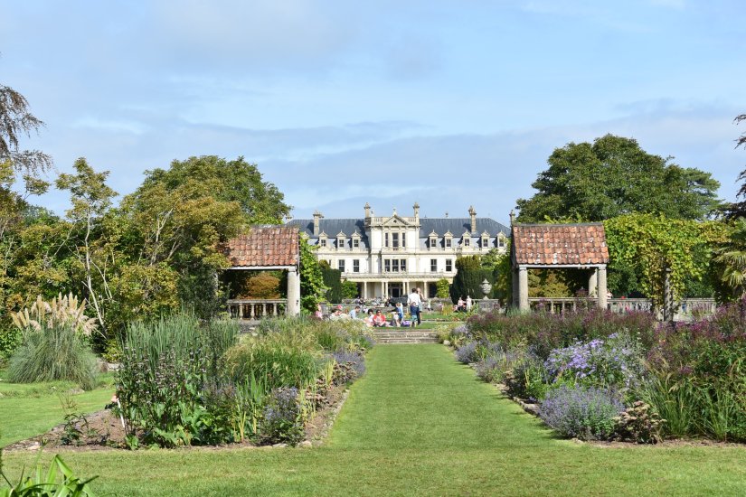A view of the grand house but further back with symmetrical lines of hedges and flower beds as far as the eye can see.
